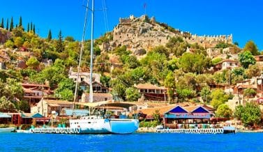 Kaş limanında demirlemiş yelkenli tekne ve arkasında tarihi kale manzarası; Dalaman Havalimanı Kaş transfer rotanızdaki eşsiz duraklar.